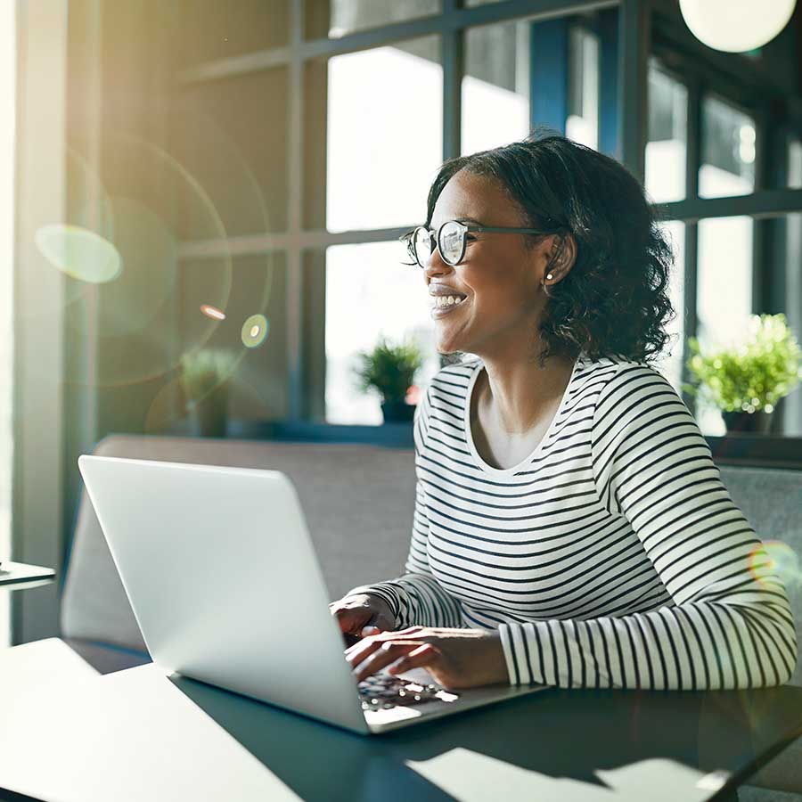 Smiling young African woman wearing glasses looking out of a window while sitting at a table working online with a laptop