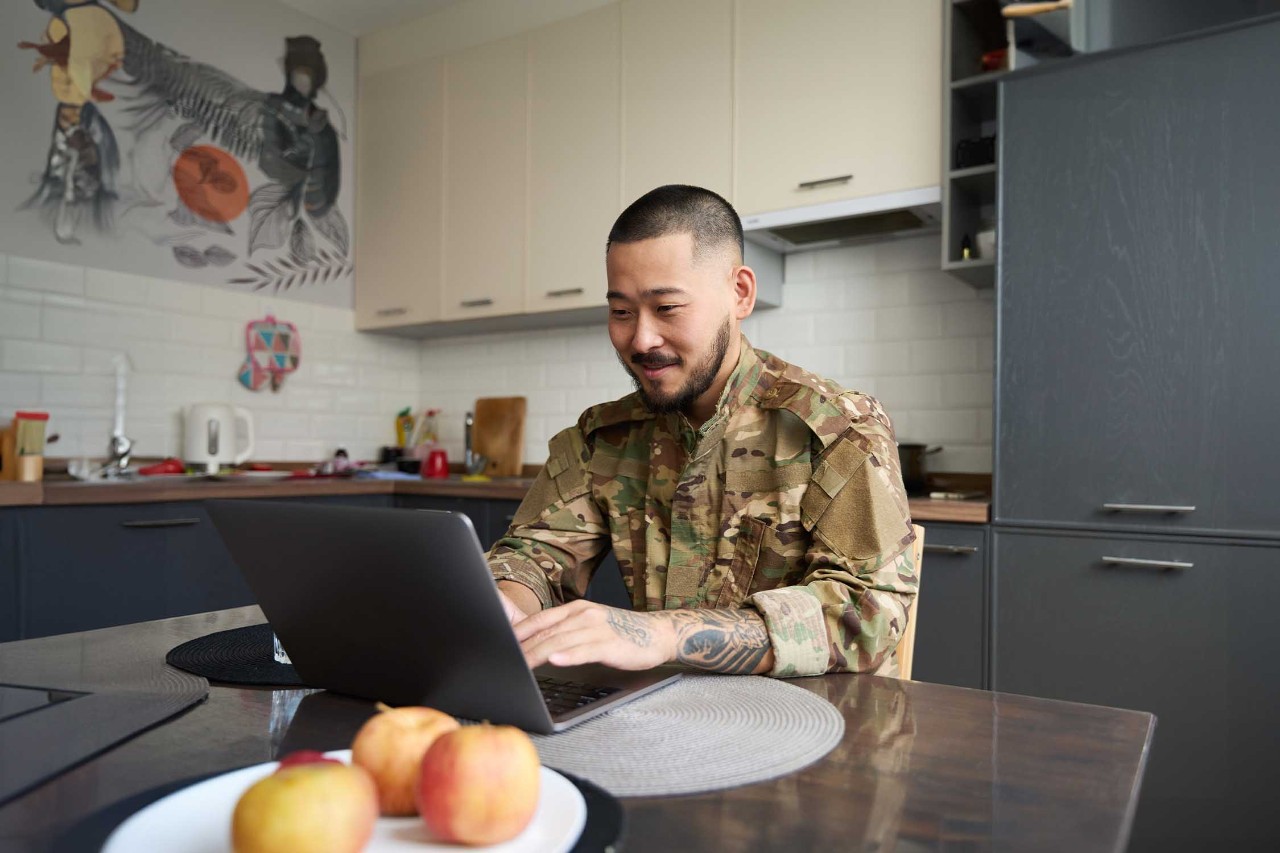 Asian male military officer in camouflage clothes sits in bright kitchen room at table and works on laptop